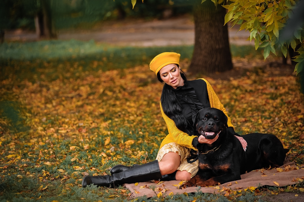 A beautiful brunette wearing an orange hat and coat sits with her well-behaved Rottweiler on a picnic blanket during a lovely autumn day.