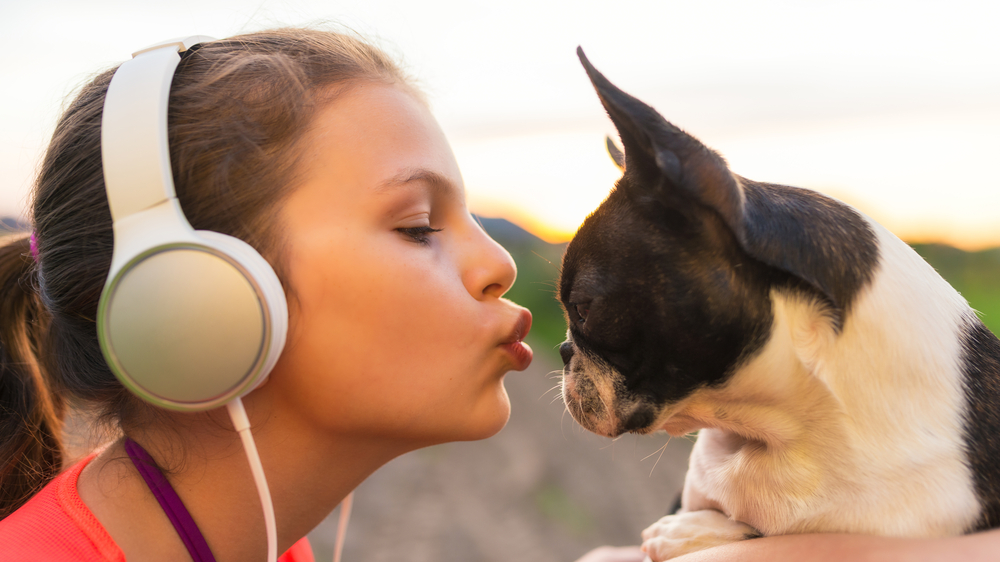 A girl wearing big headphones gives her dog a kiss while they hang out outside as the sun sets over the hills behind them.